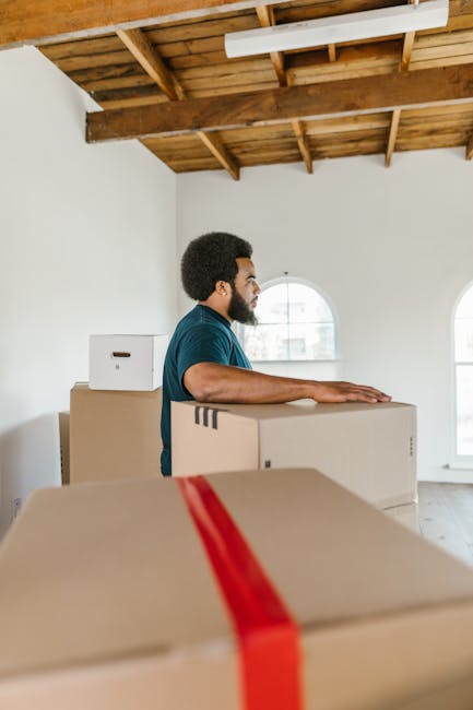 A man working on a home relocation or furniture transport process inside a property, carrying a green upholstered sofa with a tufted backrest on his shoulder, preparing to move it through an interior 