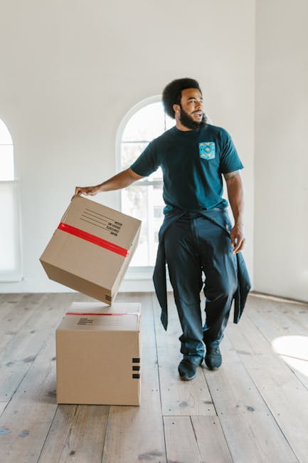 A young man wearing a blue jumpsuit and a headband stands inside a room with a light blue, textured wall. He is holding a clipboard and appears to be inspecting or organizing packing details. Surround